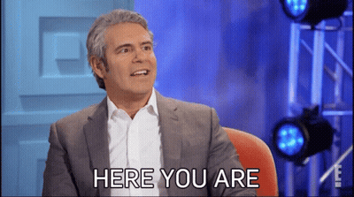 A Man In A Suit And White Shirt Is Sitting In Front Of A Sign That Says Here You Are GIF