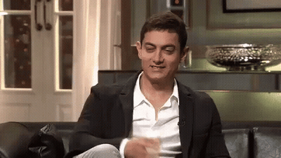 A Man In A Suit And White Shirt Is Sitting On A Couch With His Legs Crossed And Making A Funny Face . GIF