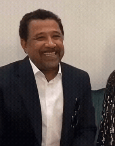 A Man In A Suit And White Shirt Is Smiling And Holding A Pair Of Glasses In His Pocket GIF