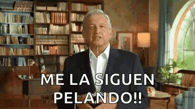 A Man In A Suit And White Shirt Is Standing In A Living Room With A Bookshelf In The Background . GIF