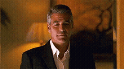 A Man In A Suit And White Shirt Is Standing In A Room And Looking At The Camera . GIF