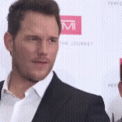 A Man In A Suit And White Shirt Is Standing On A Red Carpet And Looking At The Camera . GIF