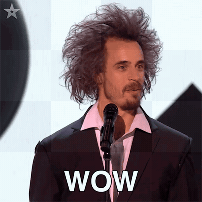 A Man In A Suit And White Shirt Stands In Front Of A Microphone With The Word Wow On His Face GIF