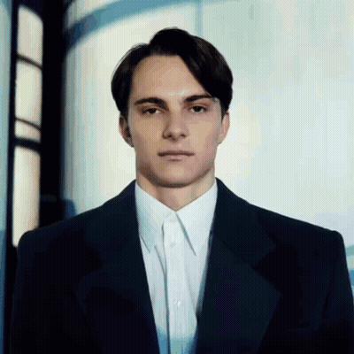 A Man In A Suit And White Shirt Stands In Front Of A Window GIF
