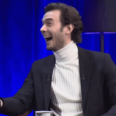 A Man In A Suit And White Turtleneck Is Laughing With His Mouth Open GIF