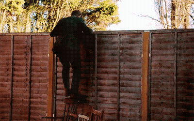 A Man In A Suit Climbs A Wooden Fence With Trees In The Background GIF