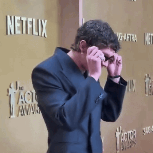 A Man In A Suit Covering His Eyes In Front Of A Netflix Sign GIF