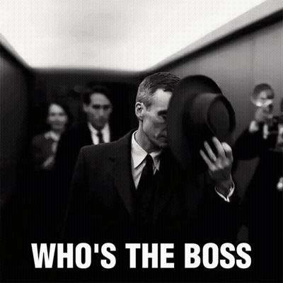 A Man In A Suit Covering His Face With His Hat And The Words Who 'S The Boss Behind Him GIF
