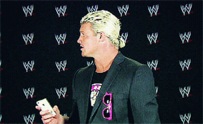 A Man In A Suit Holds A Cell Phone In Front Of A Wall With Wrestlers Logos On It GIF