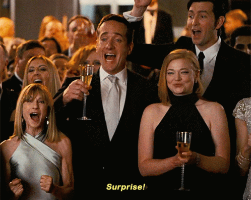 A Man In A Suit Holds A Glass Of Champagne In Front Of A Crowd That Says Surprise GIF