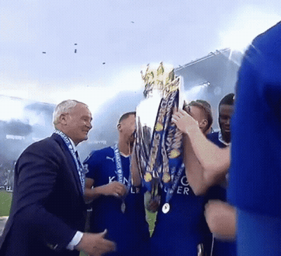 A Man In A Suit Holds A Trophy In Front Of A Group Of Soccer Players In Blue Jerseys GIF