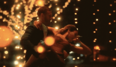 A Man In A Suit Holds A Woman In His Arms While They Dance In Front Of A String Of Lights GIF