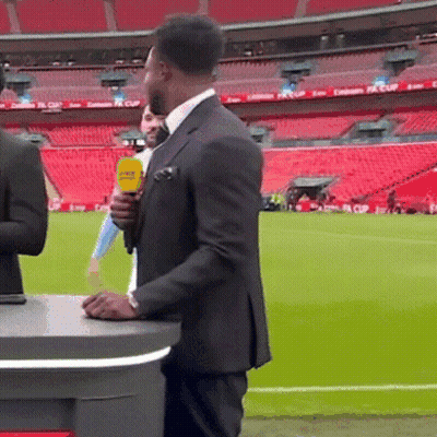 A Man In A Suit Holds A Yellow Microphone In Front Of A Stadium That Has Emirates Fa Cup Written On The Seats GIF