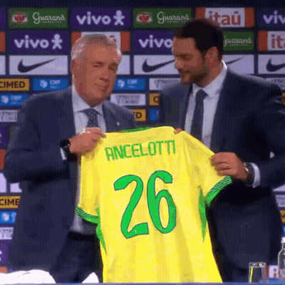 A Man In A Suit Holds Up A Yellow Shirt That Says Ancelotti 26 GIF
