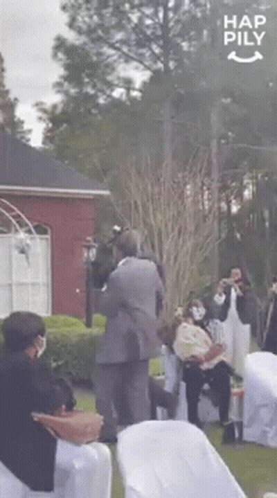 A Man In A Suit Is Carrying A Baby In His Arms While A Group Of People Are Sitting In Chairs . GIF