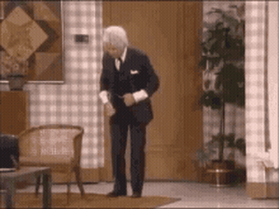 A Man In A Suit Is Dancing In A Living Room With A Chair And A Plant . GIF