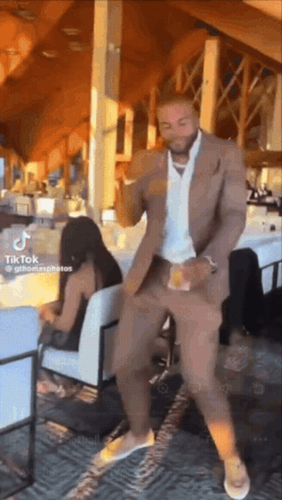 A Man In A Suit Is Dancing In A Restaurant With A Woman Sitting In A Chair Behind Him . GIF
