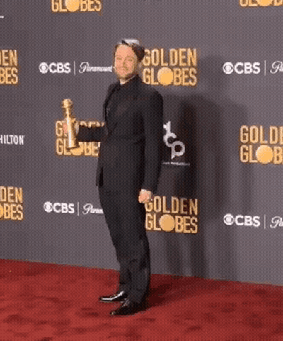 A Man In A Suit Is Dancing On A Red Carpet In Front Of A Wall That Says Golden Globes GIF