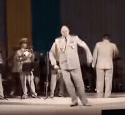 A Man In A Suit Is Dancing On A Stage While A Band Plays Behind Him . GIF
