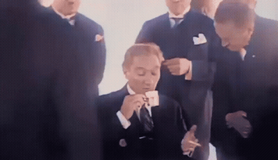 A Man In A Suit Is Drinking A Cup Of Coffee And Smoking A Cigarette While A Group Of Men Look On . GIF