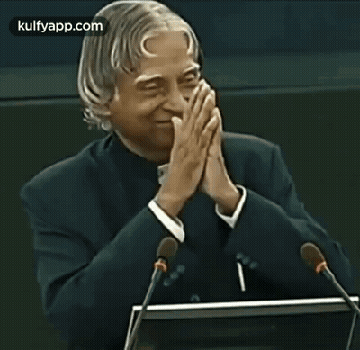 A Man In A Suit Is Giving A Speech With His Hands Folded In Front Of Two Microphones . GIF
