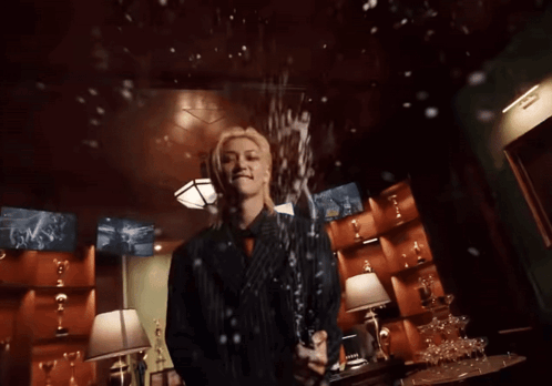 A Man In A Suit Is Holding A Bottle Of Champagne In A Room Filled With Trophies GIF