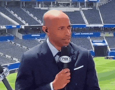 A Man In A Suit Is Holding A Fox Microphone In Front Of A Stadium . GIF
