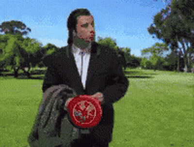 A Man In A Suit Is Holding A Frisbee In A Park With Trees In The Background GIF