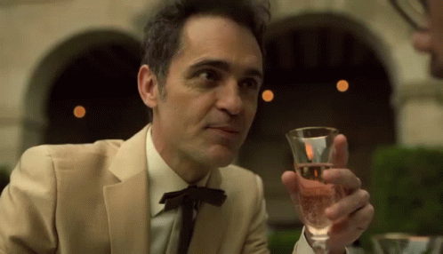 A Man In A Suit Is Holding A Glass Of Wine In His Hand . GIF