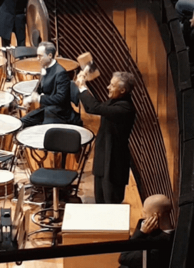 A Man In A Suit Is Holding A Hammer In His Hand In Front Of A Drum Set GIF
