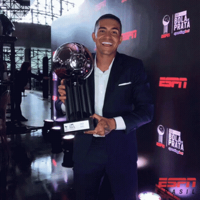 A Man In A Suit Is Holding A Trophy In Front Of A Wall That Says Bola De Prata GIF