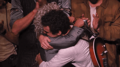 A Man In A Suit Is Hugging Another Man While Holding A Guitar GIF