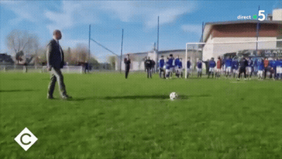 A Man In A Suit Is Kicking A Soccer Ball On A Field With A Direct 5 Logo In The Corner GIF