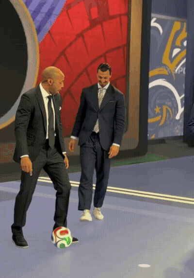 A Man In A Suit Is Kicking A Soccer Ball While Another Man Watches GIF