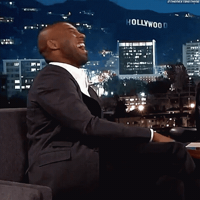 A Man In A Suit Is Laughing In Front Of A Sign That Says Hollywood GIF