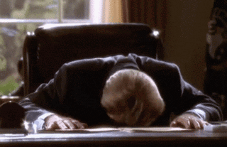 A Man In A Suit Is Laying His Head On A Desk In Front Of A Window GIF