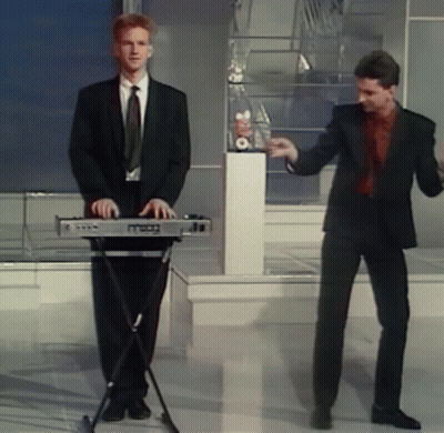 A Man In A Suit Is Playing A Keyboard While Another Man Dances Behind Him GIF