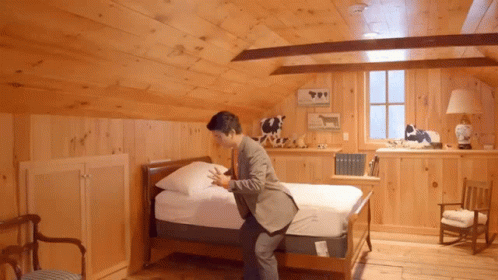 A Man In A Suit Is Putting A Mattress On A Bed In A Wooden Room GIF