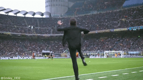 A Man In A Suit Is Running On A Soccer Field In Front Of A Stadium That Says Ambrosiaba GIF