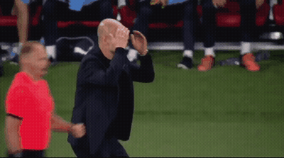 A Man In A Suit Is Running On A Soccer Field With A Referee In A Red Shirt Behind Him GIF