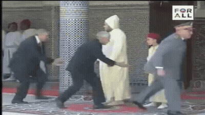 A Man In A Suit Is Shaking Hands With A Man In A White Robe While Walking Down A Red Carpet . GIF