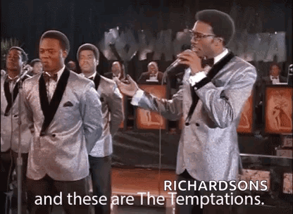 A Man In A Suit Is Singing Into A Microphone In Front Of A Group Of Men In Suits . GIF