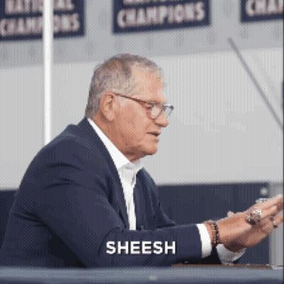 A Man In A Suit Is Sitting At A Table And The Word Sheesh Is On His Arm GIF
