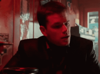A Man In A Suit Is Sitting At A Table In A Dark Room With A Red Light Behind Him . GIF