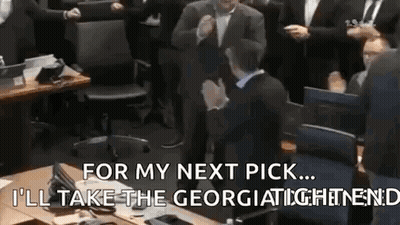 A Man In A Suit Is Sitting At A Table In Front Of A Crowd Of People And Talking To Them . GIF