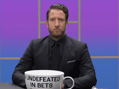 A Man In A Suit Is Sitting At A Table With A Cup That Says " Undefeated In Bets " GIF