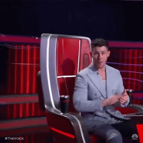 A Man In A Suit Is Sitting In A Chair On A Stage With A Red Curtain In The Background . GIF