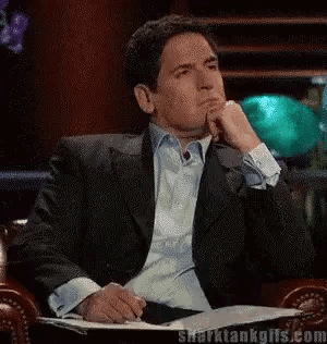 A Man In A Suit Is Sitting In A Chair With His Hand On His Chin And A Book In Front Of Him . GIF
