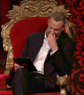 A Man In A Suit Is Sitting In A Red Chair Holding A Tablet And Covering His Mouth GIF