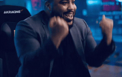 A Man In A Suit Is Sitting In An Akracing Chair With His Arms In The Air GIF